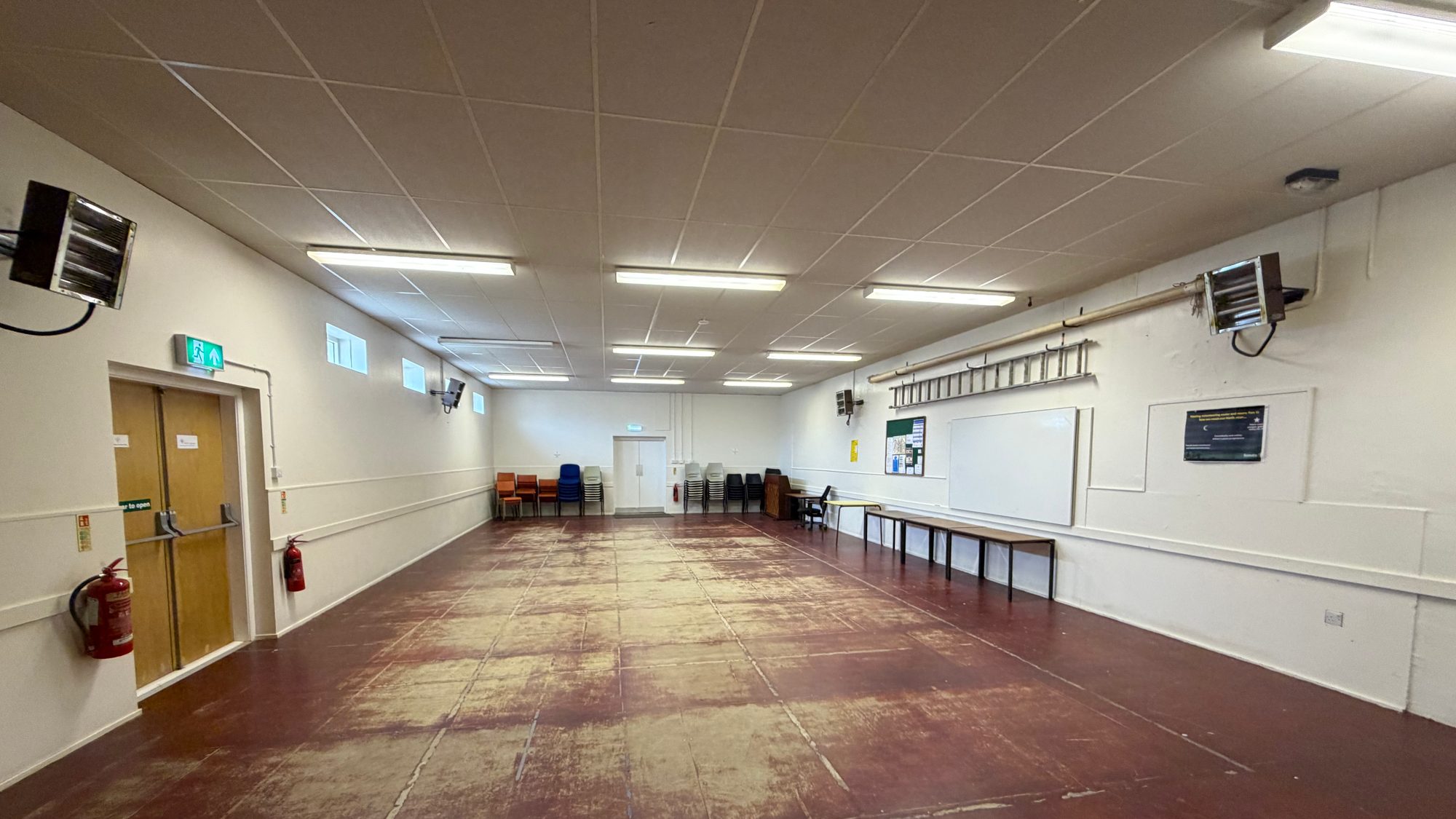 The main hall at Dalneigh Scout Hall, a long open room seen from one end, with a fire door on the left and a whiteboard along the right-hand wall.