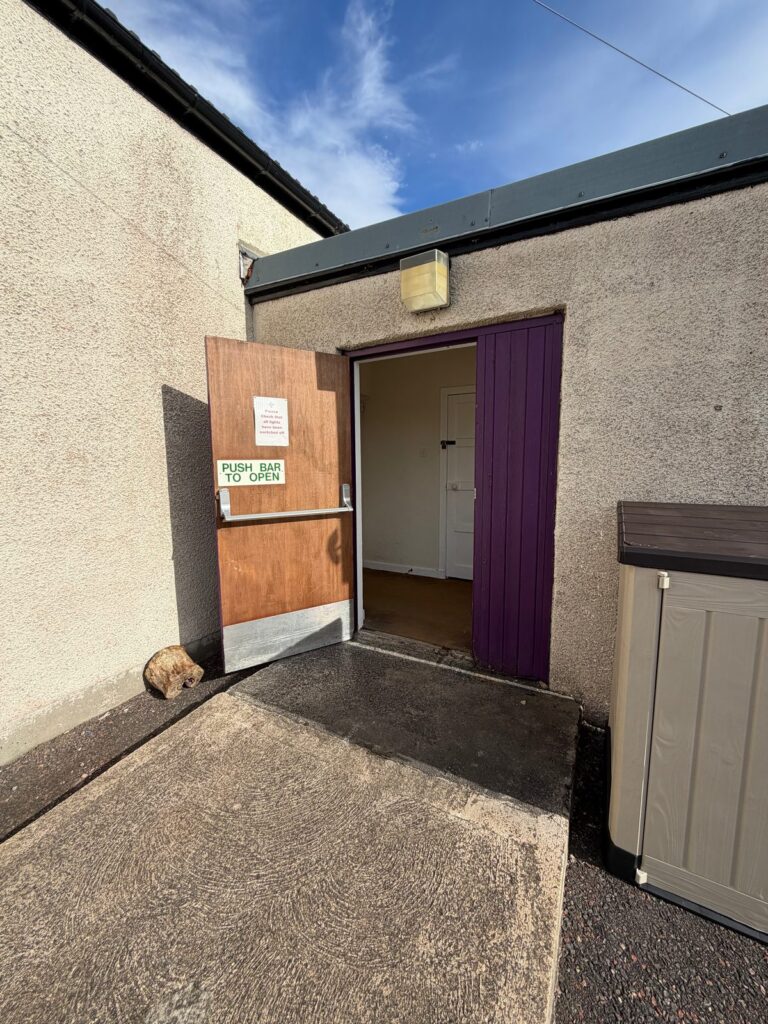 The side door to the main hall from the car park, with a ramp up to the door and a storage box beside it.